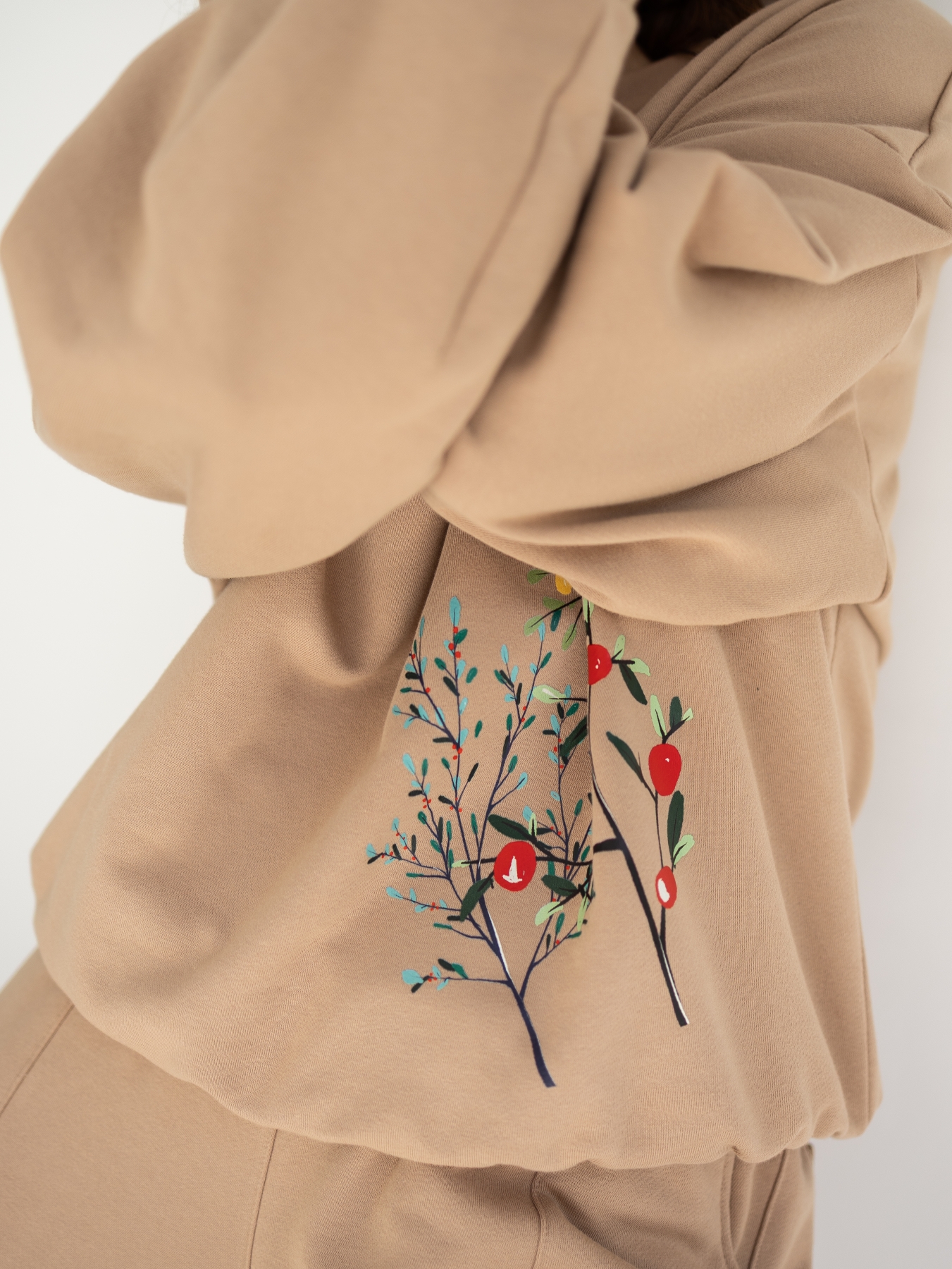 Beżowa bluza oversize z długim rękawem i subtelnym roślinnym printem z przodu, zestawiona z beżowymi spodniami. Modelka stoi przy jasnej ścianie.