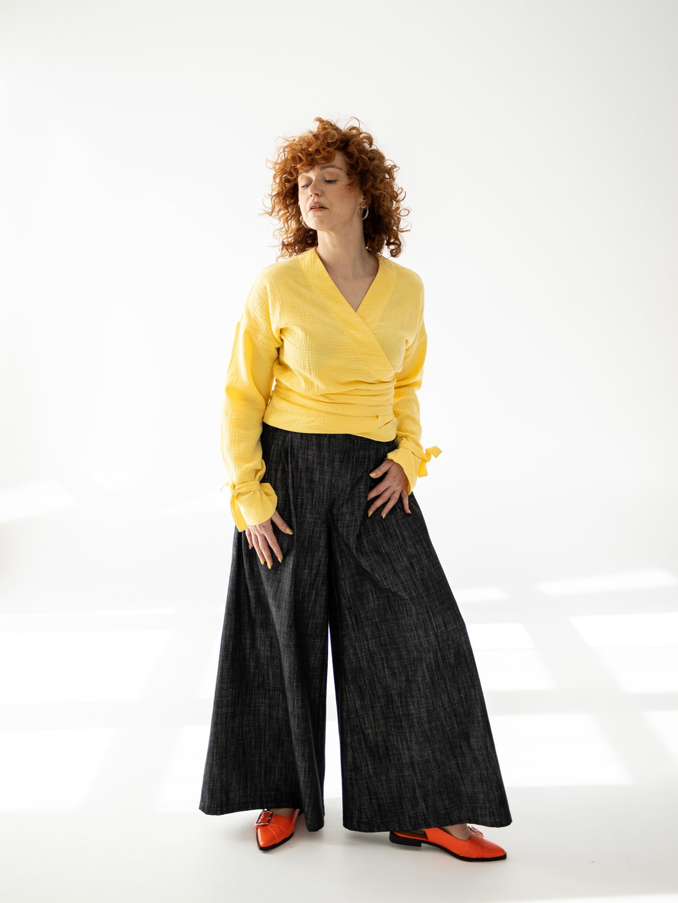 Modelka w szerokich, ciemnych bawełnianych spodniach z wysokim stanem i bardzo szeroką nogawką, zestawionych z żółtą kopertową bluzką.