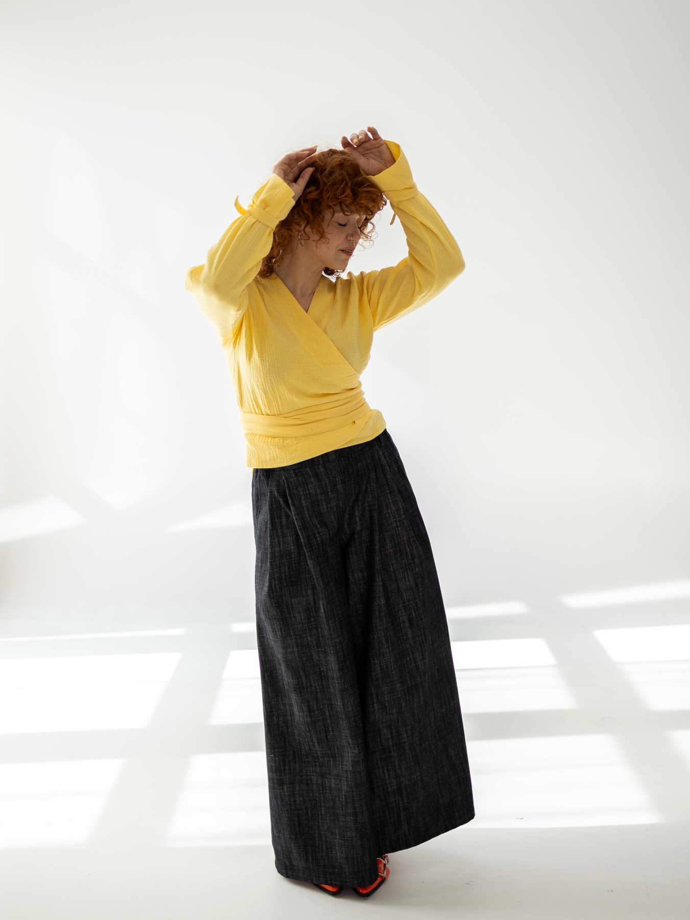 Modelka w szerokich, ciemnych bawełnianych spodniach z wysokim stanem i bardzo szeroką nogawką, zestawionych z żółtą kopertową bluzką.