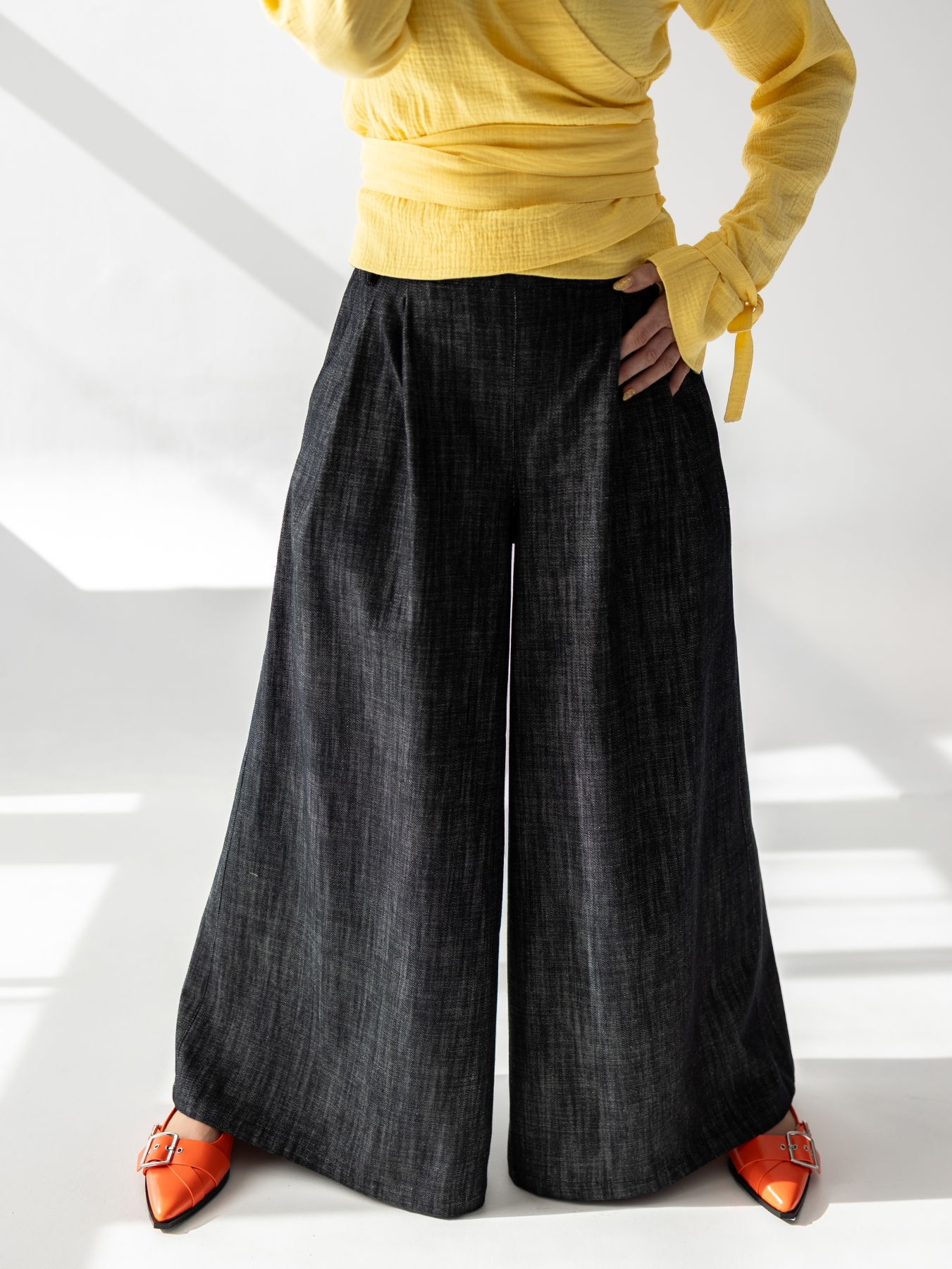 Modelka w szerokich, ciemnych bawełnianych spodniach z wysokim stanem i bardzo szeroką nogawką, zestawionych z żółtą kopertową bluzką.